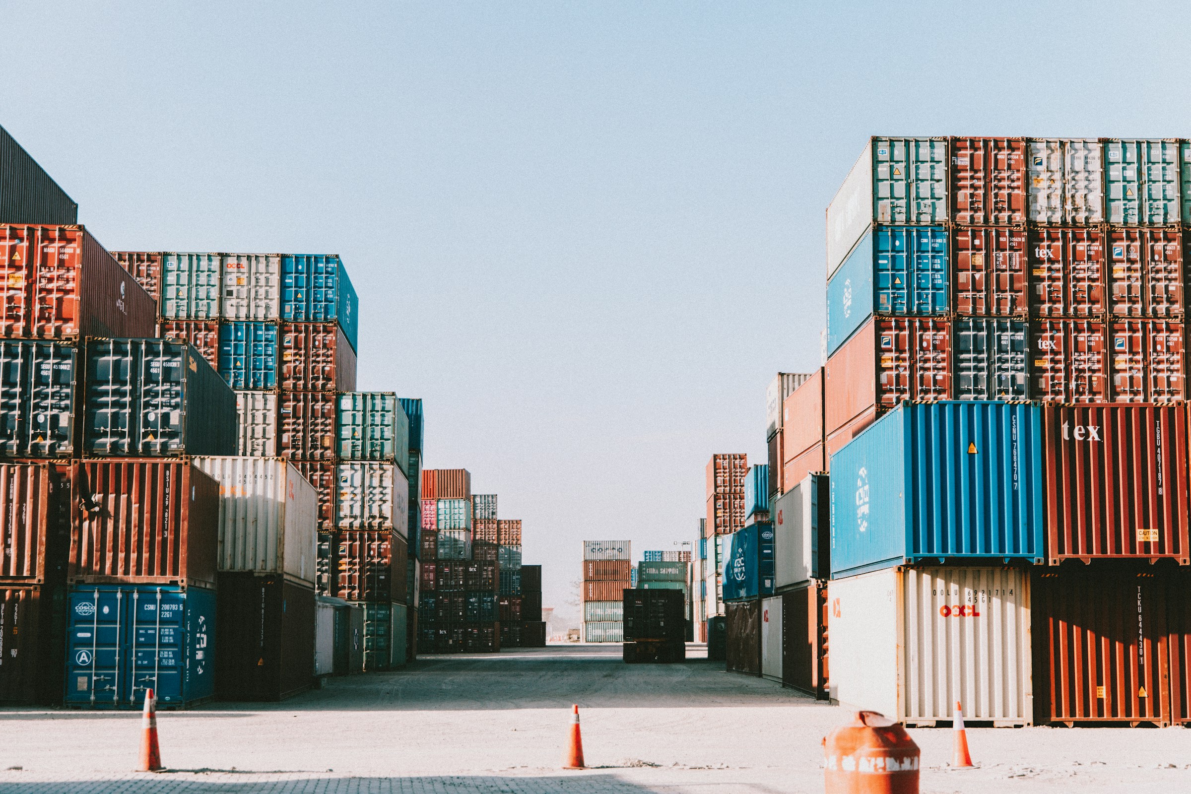 Tall stacks of weathered shipping containers line an empty lane in a port yard under a pale sky.